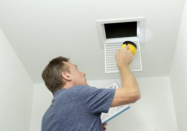 Man inspecting air ducts shining a flashlight through a small square ceiling vent into ducting pipes. Mature male examining the condition of air ducts at home.